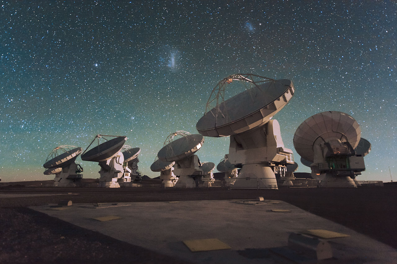 Antennas of the Atacama Large Millimeter/submillimeter Arrangement (ALMA), on the Chajnantor Plateau in the Chilean Andes. The Large and Small Magellanic Clouds, two accompaniment galaxies to our own Milky Way galaxy, can be apparent as ablaze smudges in the night sky, in the centre of the photograph. ALMA ATTACAMA