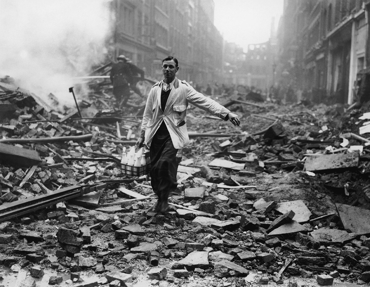 A milkman delivers milk in a London street devastated during a German bombing raid.  Firemen are dampening down the ruins behind him