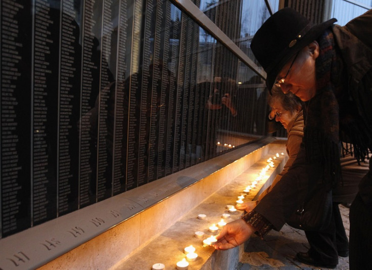 People light candles in front of a wall containing the names of Holocaust victims