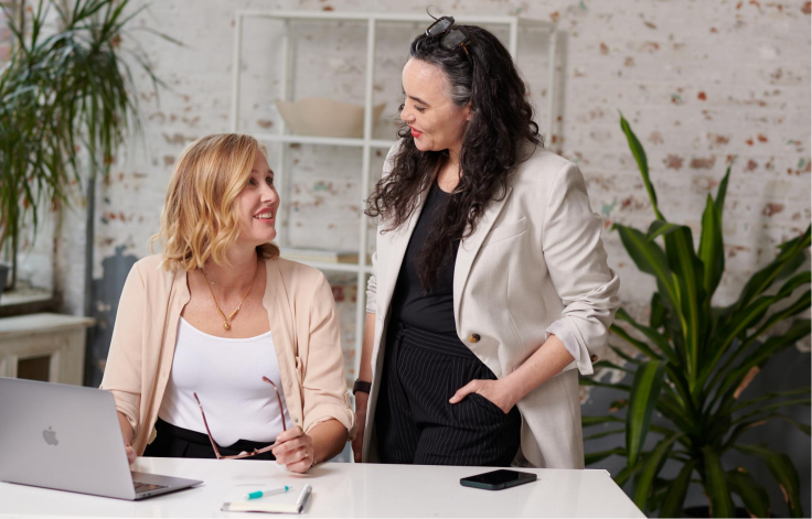 Two women chat comfortably in the office sporting Become’s signature 