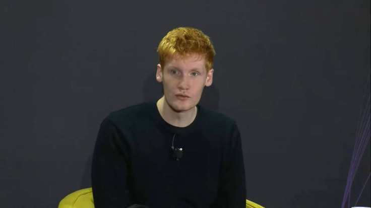 25-year-old red-haired man sitting in white armchair on stage, speaking