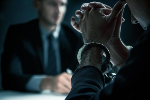 A man in handcuffs sits across from another man in a suit during an interrogation