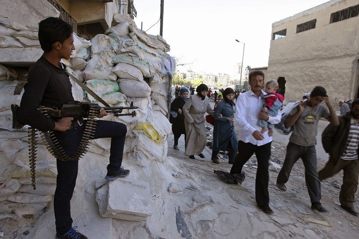 A Free Syrian Army fighter carries his weapon and stands guard as civilians walk past.